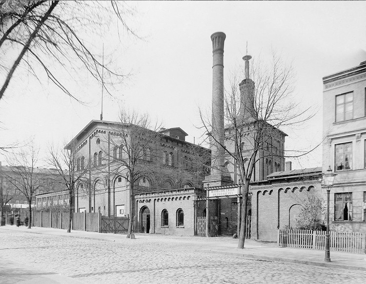 Schwarz-weiß Aufnahme einer Fabrik mit davorliegender Allee.